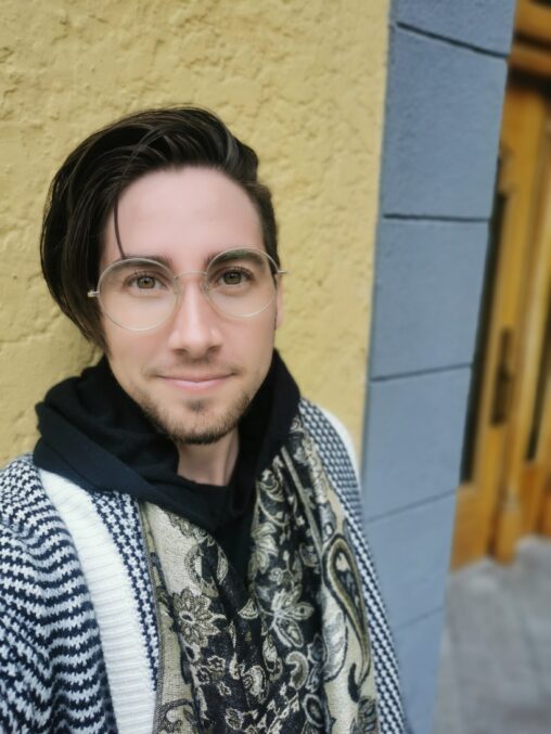 a white skinned person wearing round, gold rimmed glasses, smiles into the camera. their hair is dark brown and shaved on one side, and about chin length on the other. They also have some dark facial hair on their chin. they are wearing a black and white checked sweater with a black and gold scarf.